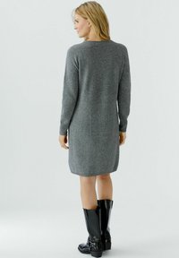Robe-pull en tricot gris avec des manches longues, des poignets côtelés et une silhouette droite, associée à des bottes noires laquées jusqu'au genou.
