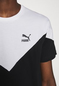 Detail černobílého trička Puma s logem na horní části hrudi, nošeného osobou s tmavou barvou pleti.