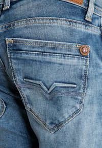 El bolsillo trasero de unos jeans de mezclilla azul con costuras decorativas y un pequeño detalle de botón de cuero redondo cerca de la esquina superior derecha.