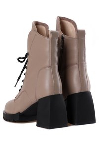 Stivaletti alla caviglia in pelle beige con un alto tacco a blocco nero. Le caratteristiche includono lacci frontali, cerniera laterale e colletto imbottito. Design strutturato.