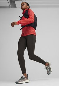 Rote leichte Jacke über einem schwarzen Weste, kombiniert mit braunen Leggings und gemusterten Laufschuhen. Der Hintergrund ist schlicht weiß, was das Outfit hervorhebt.