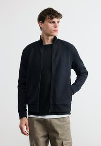 Νεαρός άνδρας που φοράει μαύρο μπουφάν με φερμουάρ, μαύρο πουκάμισο και μπεζ cargo παντελόνια, κοιτάζει προς το πλάι με το ένα του χέρι στην τσέπη.