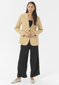 Blazer beige con un unico bottone nero, maniche lunghe e vestibilità su misura, abbinato a pantaloni neri a gamba larga e ballerine di colore chiaro.