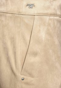 Beige samettihousut, joissa on ommeltu etutasku, hopeanväriset metalliosat ja brändin logo-tunnus taskun yläpuolella.