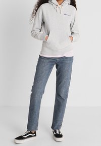 Gray hoodie with a front pocket, drawstrings, and purple branding; paired with light blue jeans and black and white sneakers.