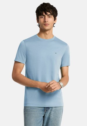 Giovane uomo con capelli scuri che indossa una maglietta azzurro chiaro a maniche corte e jeans, in piedi con le mani giunte davanti.