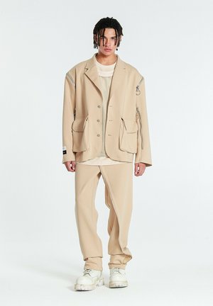 Joven con el pelo trenzado que lleva una chaqueta beige oversize con grandes bolsillos, pantalones a juego y zapatos blancos gruesos, de pie frente a un fondo blanco.
