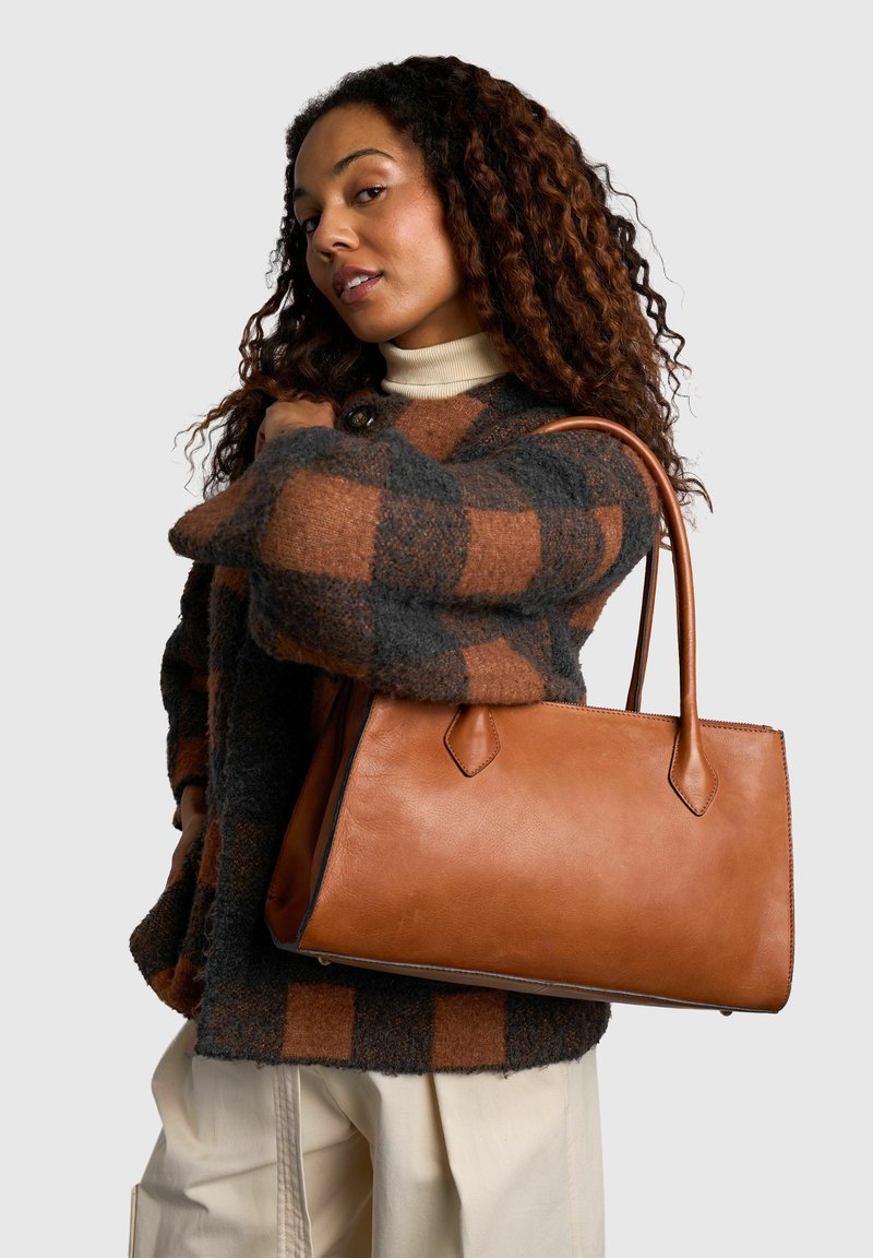 Femme aux cheveux bouclés portant une veste à carreaux marron et grise, portant un grand sac à main en cuir beige sur l’épaule sur un fond neutre.