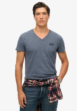 Camiseta de algodón azul jaspeado con escote en V y mangas cortas, combinada con una camisa de franela a cuadros atada a la cintura, y un reloj plateado en la muñeca.