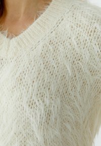 Close-up van een crèmekleurige, pluizige, gebreide trui met V-hals gedragen door een persoon, met de nadruk op de zachte textuur en het losse breipatroon.