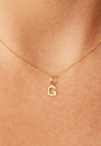 Collier en or avec une chaîne fine et délicate, ornée d'un pendentif en forme de lettre "g" minuscule. Le métal a une finition polie qui reflète la lumière subtilement.