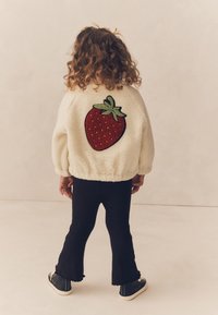 Hvid fleecejakke med en stor rød jordbærbrodering på ryggen, kombineret med sorte udvidede bukser og sorte sneakers.