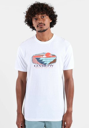T-shirt en coton blanc avec un graphisme coloré représentant un coucher de soleil et des vagues. Le design inclut le texte "OXBOW" avec des accents bleus et rouges.