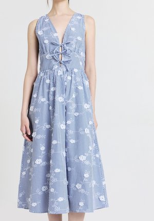 Frau trägt ärmelloses blau-weiß gestreiftes Kleid mit weißer Blumenstickerei und gekreuzter Schnürung vorne.