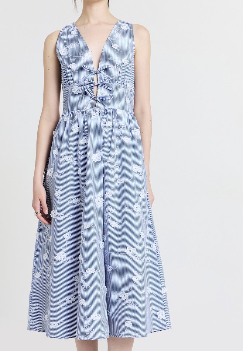 Femme portant une robe sans manches rayée bleu et blanc avec broderie florale blanche et détail lacé croisé à l'avant.