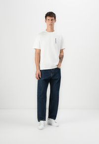 White cotton t-shirt with short sleeves, featuring a small blue logo on the left chest; paired with dark blue straight-leg jeans and white sneakers.