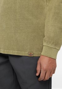 Long-sleeve olive green shirt made of soft fabric, featuring a ribbed collar, and a small logo patch near the bottom seam.