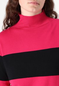 Pink and black striped sweater with a high collar, made of a soft knit fabric. Features a bold horizontal black stripe against a pink background.