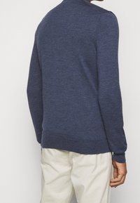 Pull en tricot bleu foncé avec un col rond, des poignets et un ourlet côtelés, et une finition texturée. Porté avec un pantalon de couleur claire.