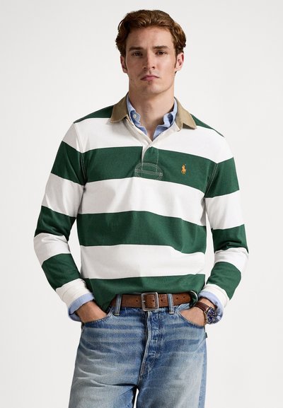 Vīrietis, kurš valkā zaļu un baltu plaši svītrainu regbija kreklu pār gaiši zilu pogājamu kreklu, zilas džinsas, brūnu jostu un rokas pulksteni, stāv ar rokām kabatās.