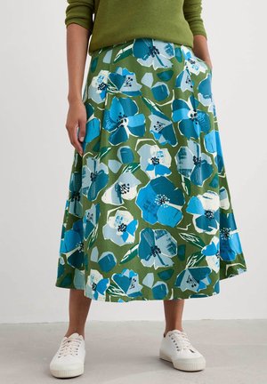 Personne portant une jupe verte à grands motifs floraux bleus et blancs, un pull vert côtelé, des baskets blanches, debout les mains dans les poches.