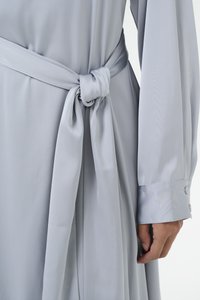 Robe longue gris clair en tissu lisse, avec une ceinture nouée à la taille et des poignets boutonnés. La texture semble douce et fluide.