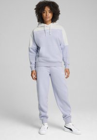 Felpa viola chiaro con accenti bianchi e logo, abbinata a pantaloni della tuta coordinati. Texture morbida, vestibilità comoda, con tasca frontale. Sneakers bianche.