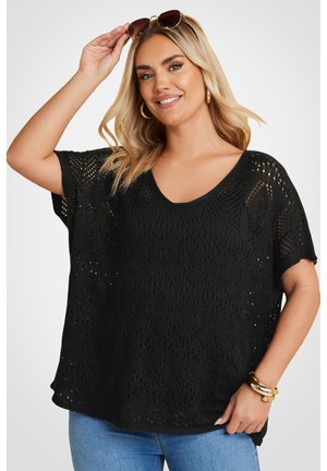 Jersey negro de punto con corte holgado, escote en V, mangas cortas y patrón de tejido abierto. Combinado con jeans de mezcla clara y pulseras doradas.