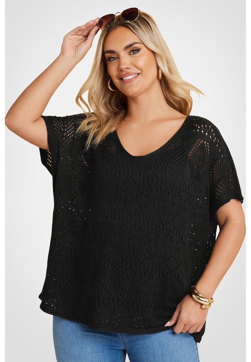 Jersey negro de punto con corte holgado, escote en V, mangas cortas y patrón de tejido abierto. Combinado con jeans de mezcla clara y pulseras doradas.