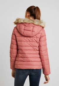 Lyserød puffjakke med hætte i faux pels. Har horisontale quiltede sektioner, lange ærmer og elastiske manchetter. Bagrenden viser en tætsiddende silhuet.
