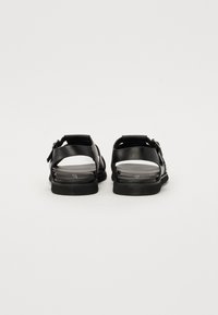 Black sandals with two adjustable straps, a flat sole, and a smooth texture. Features metallic buckle accents and open-back design.