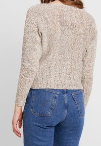 Beżowy sweter dziany z wielokolorowymi plamkami, z długimi rękawami i skróconym dołem. Połączony z niebieskimi dżinsami.