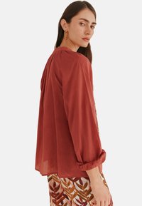 Rostfarbene, locker sitzende Bluse mit langen Ärmeln. Verfügt über eine geraffte Detail am Rücken und weichen Stoff. Kombiniert mit einem gemusterten Blätterrock.