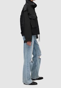 Zwarte, oversized jas met zakken en hoge kraag; gecombineerd met vervaagde blauwe, wijd uitlopende gescheurde spijkerbroeken en zwarte instapschoenen.