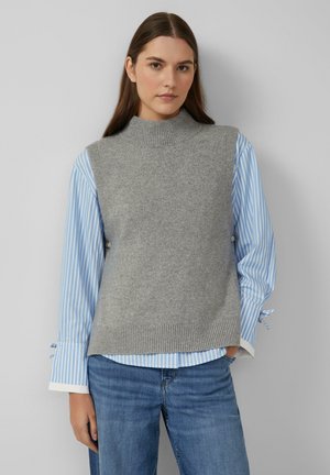 Vrouw met lang bruin haar, gekleed in een grijze mouwloze gebreide vest over een blauw-wit gestreept shirt en spijkerbroek, staande tegen een effen achtergrond.
