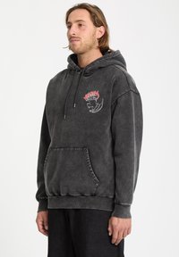 Szary hoodie z regulowanym sznurkiem, kieszenią z przodu oraz haftowanym wzorem w czerwonym i białym kolorze z motywem smoka. Wykonany z miękkiej tkaniny.