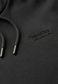 Hoodie noir en tissu doux, avec un logo sur le devant en lettres embossées et des cordons de serrage avec des embouts en métal pour l'ajustement.