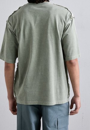 Olivgrünes Oversize-T-Shirt aus weicher Baumwolle mit rohen Kanten und seitlichen Schlitzen. Kurze Ärmel und ein rundes Halsbündchen.