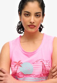 Jonge vrouw met donker haar, gekleed in een roze mouwloos shirt met een roodblauw tropisch eiland- en zeilbootmotief, met gekruiste armen.