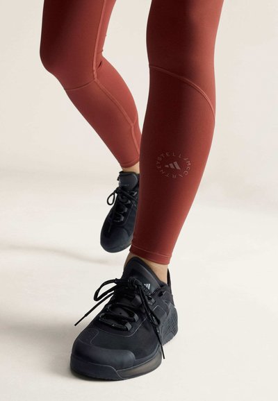 Person trägt rostfarbene Leggings und schwarze Sportschuhe und steht mit einem leicht angehobenen Fuß.