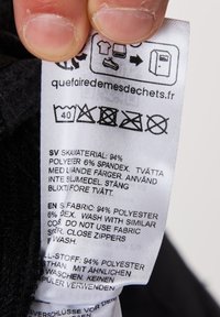 Pflegeetikett für Kleidung mit Waschhinweisen und Stoffzusammensetzung: 94 % Polyester, 6 % Spandex. Etiketten in mehreren Sprachen, weiß auf schwarz.