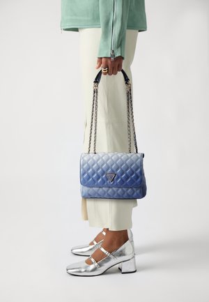 Osoba trzymająca niebieską pikowaną torebkę na łańcuszkach, ubrana w jasne spodnie, zieloną kurtkę i metaliczne srebrne buty Mary Jane.