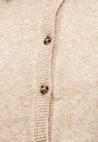 Εγγύς λήψη μπεζ πλεκτής ζακέτας με δύο κουμπιά από χελώνια που είναι κουμπωμένα κατά μήκος μιας ριγωτής λωρίδας.