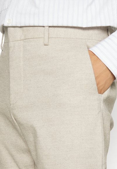 Helle beigefarbene Hose aus strukturiertem Stoff, mit seitlichen Taschen und Gürtelschlaufen. Kombiniert mit einem weißen gestreiften Hemd.