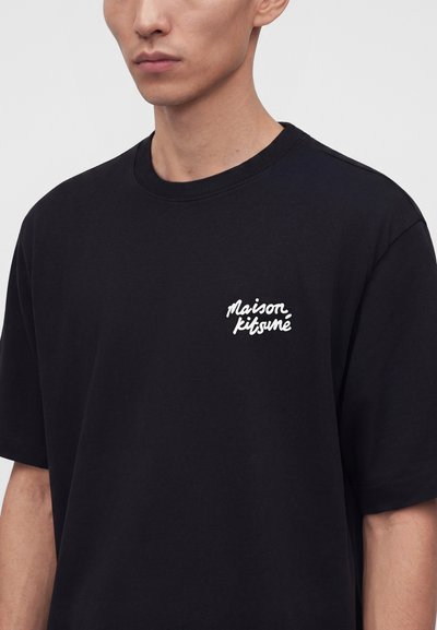 Άνδρας που φοράει ένα μαύρο κοντομάνικο t-shirt με λευκό κείμενο "Maison Kitsuné" στο αριστερό μέρος του στήθους, εμφανιζόμενος από τους ώμους μέχρι τη μέση του κορμού.