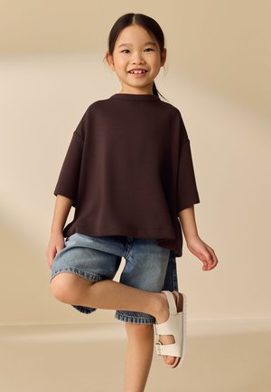 Junges Mädchen lächelt, trägt ein lockeres dunkelbraunes Hemd, Jeansshorts und weiße Sandalen, steht mit einem Bein angehoben vor einem einfarbigen beigen Hintergrund.