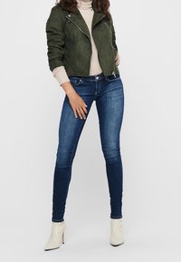 Veste en daim kaki avec fermeture éclair à l'avant, associée à un jean skinny bleu foncé et des bottines crème. Texture lisse, design ajusté.