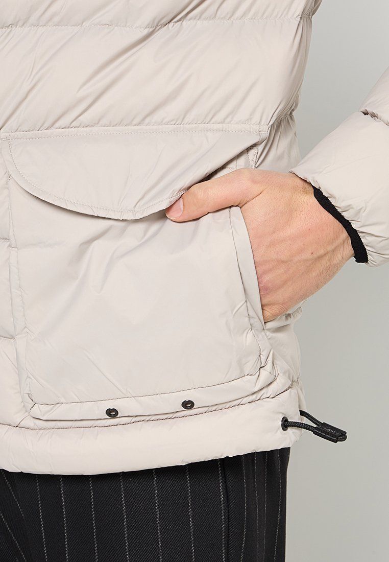 Giubbino puffer beige chiaro con texture trapuntata, tasca frontale per le mani e orlo regolabile con stringhe. Presenta dettagli neri nei polsini.