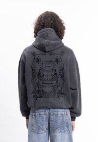 Tmavě šedá mikina s kapucí s texturovanou grafikou na zádech, která obsahuje okrasné designové prvky. Překrývající se vrstvy dodávají hloubku.
