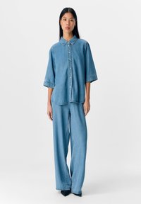 Jeune femme debout, portant une chemise en denim ample bleu clair à boutons et un jean large assorti, sur un fond blanc uni.